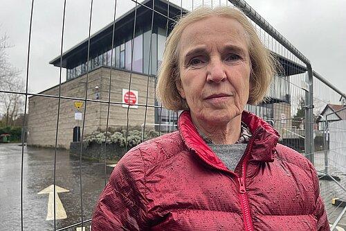 Susan Murray stands Infront of a fence in a red jacket looking serious