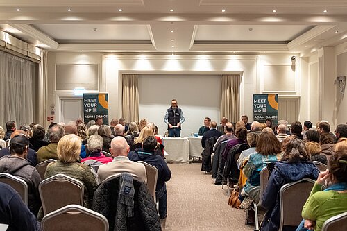 Adam Dance MP stands at the front of a packed meeting room, addressing attendees at his flooding forum. He wears a navy blue vest over a light blue shirt and speaks with his hands clasped. Behind him, a panel of speakers sits at a long table covered in a white cloth. Two large banners on either side of the stage display "MEET YOUR MP" and "ADAM DANCE." The audience, consisting of a diverse group of people, sits in rows of chairs facing the front, with some wearing coats and scarves.