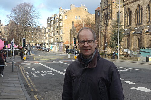 Neil Ross standing in front of Holy Corner, a junction in Morningside, with churches and tenements in the background. The sky is blue.