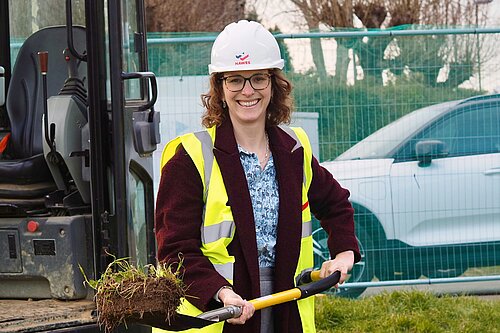 Alison Bennett MP holding a spade with a turf next to a digger near haras fencing