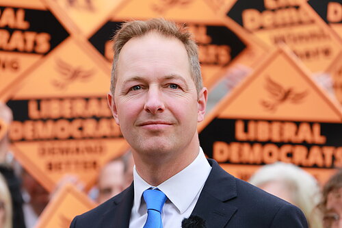 Head and shoulders image of Richard Foord in front of activists holding signs reading "Liberal Democrats winning here"