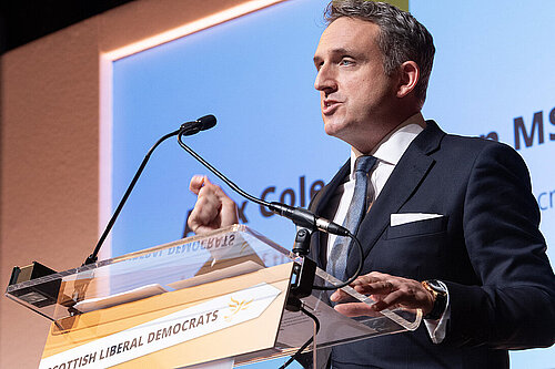 Alex Cole-Hamilton speaking at a podium with the words "Scottish Liberal Democrats" on it